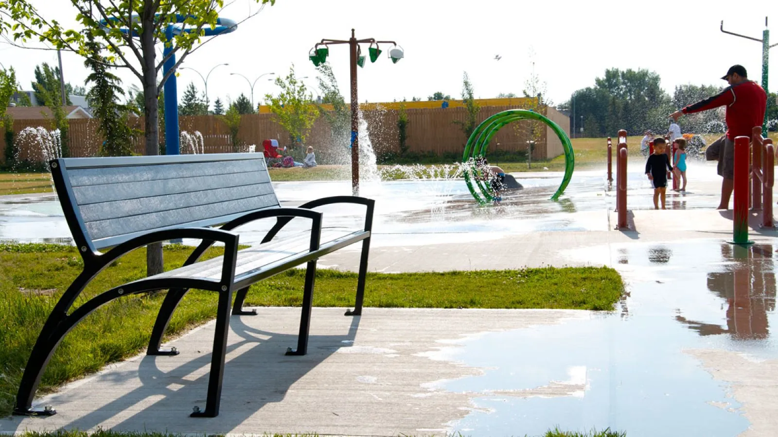 A Modena bench provides seating at the Callingwood Spray Park in Edmonton, Alberta. This park bench offers a place to rest near the splash pad.