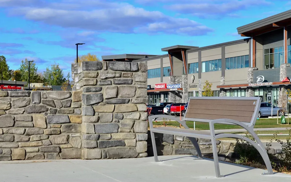 A Modena bench sits beside a stone wall in a commercial area. The bench features recycled plastic lumber and a powder-coated metal frame.