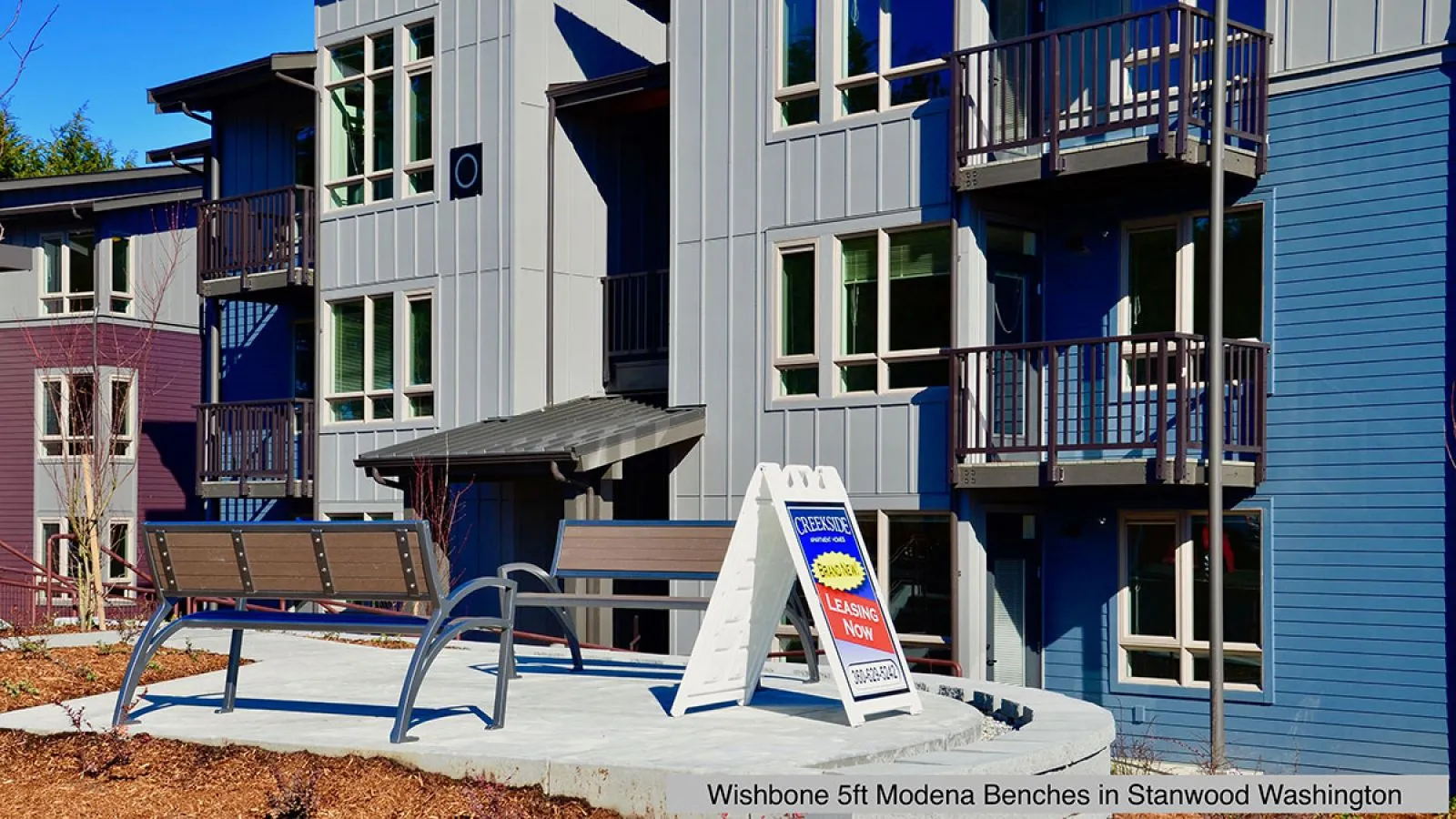 Two Modena benches provide seating outside a modern apartment complex. The benches feature recycled plastic lumber seats and metal frames.