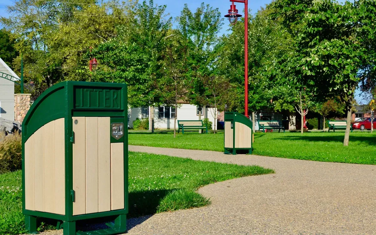 Modena curved top waste receptacles are placed along a paved path in a park-like setting. These durable litter bins feature a green frame and beige recycled plastic lumber.