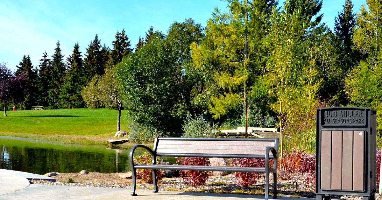 A Mountain Classic bench and Beselt Square Top Waste Receptacle are situated near a pond in Bud Miller All Seasons Park. The bench features recycled plastic lumber and a powder-coated metal frame.
