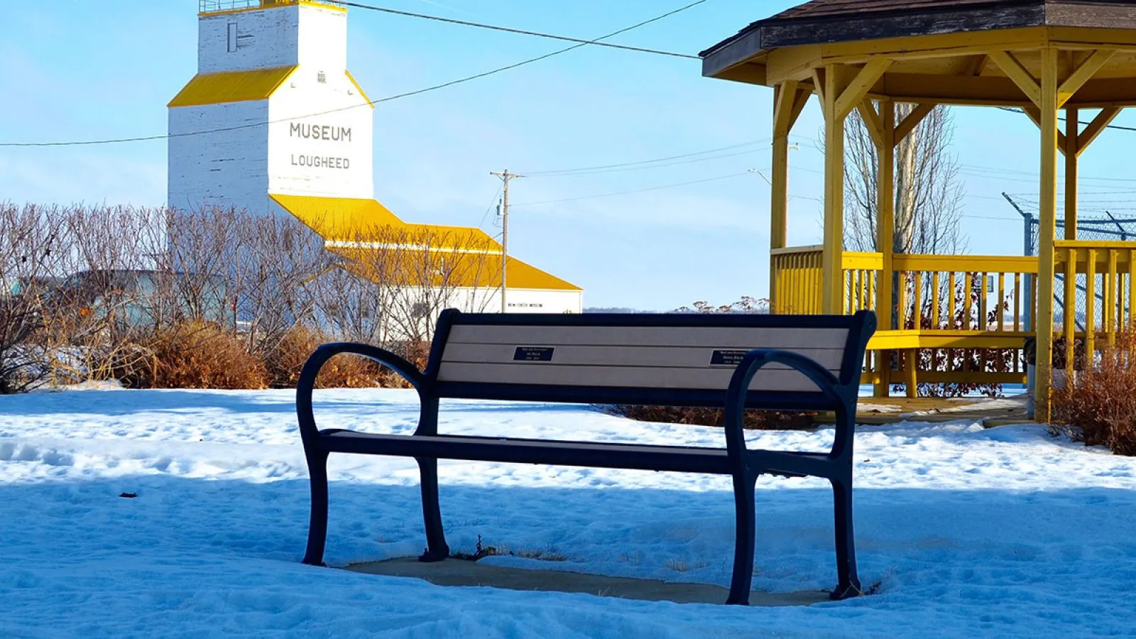 A Mountain Classic bench sits in a snow-covered park near the Lougheed Museum and a yellow gazebo. This bench provides a place to rest and enjoy the winter scenery.