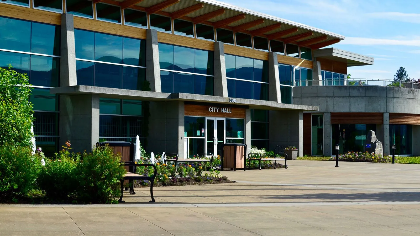 Benches and waste receptacles provide seating and waste disposal at the entrance to City Hall. The site furniture complements the building's modern architecture and landscaping.