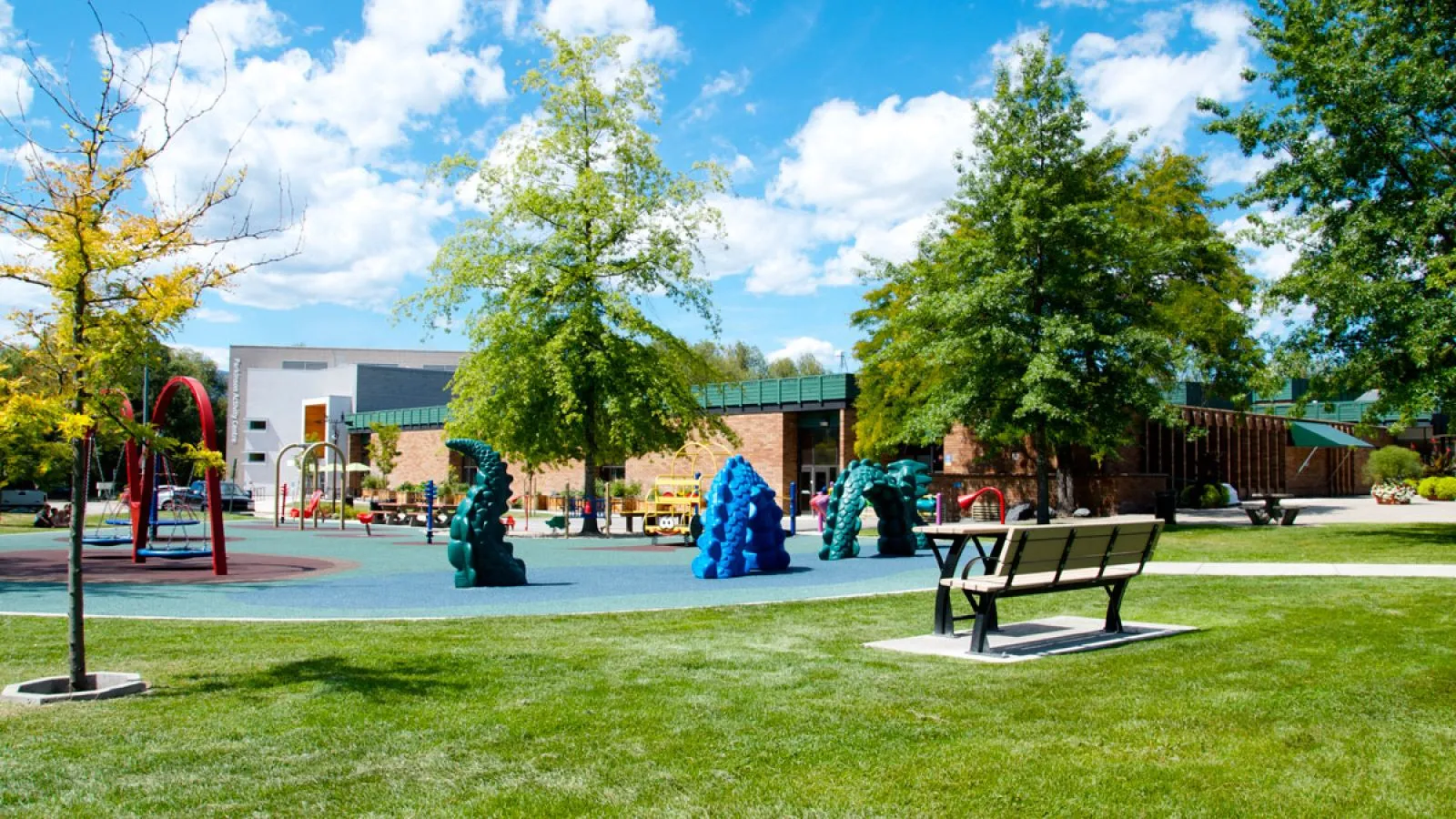 A Parker Bench and Table Combo sits on a grassy lawn near a playground. This outdoor furniture set provides a comfortable space for relaxation and socializing in a recreational area.