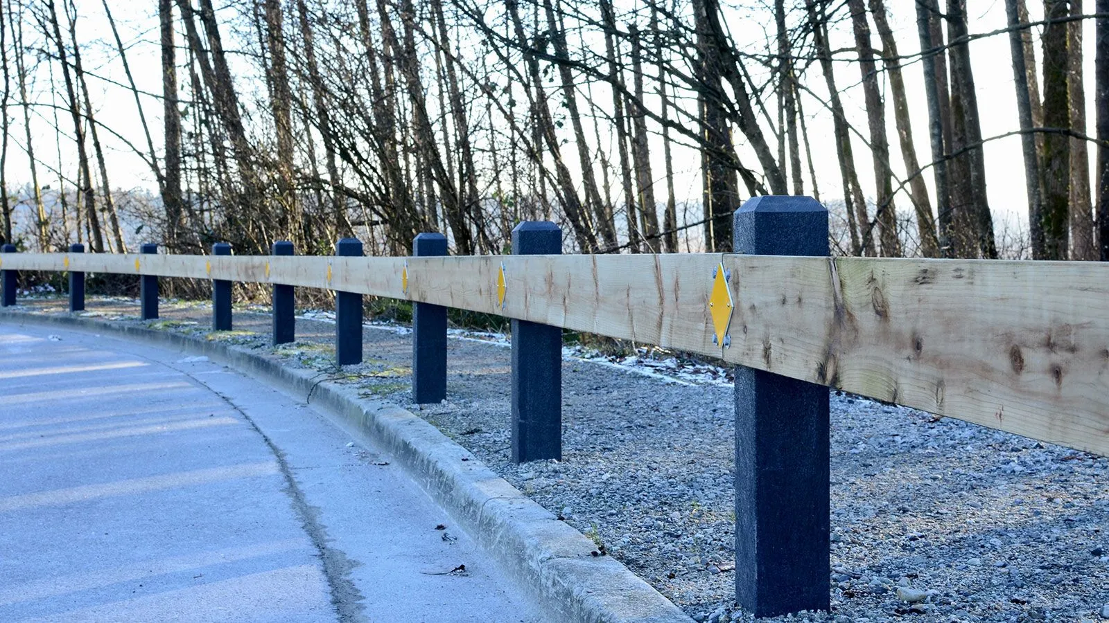 Recycled plastic bollards with a timber rail create a barrier along a roadside. The posts feature a pyramid top and are installed near a gravel shoulder and tree line in Port Coquitlam, BC.