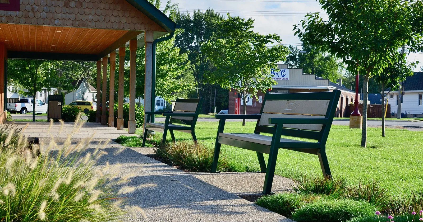 Two Rutherford Angled Leg benches provide seating in a park setting. The benches feature green frames and light-colored recycled plastic lumber.