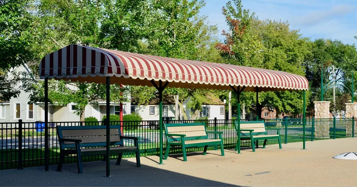 Three benches sit beneath a striped awning, providing shaded seating in a public space. The benches feature recycled plastic lumber seats and green powder-coated frames.