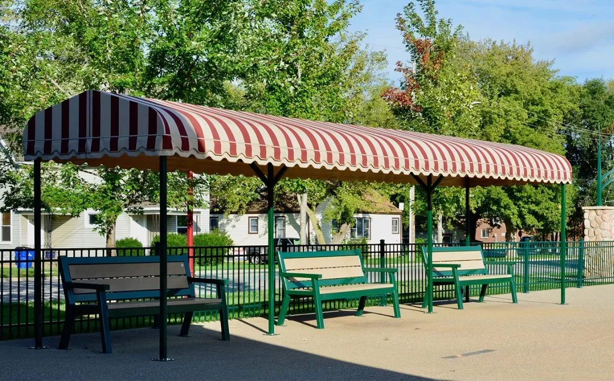 These Rutherford benches provide comfortable seating beneath a striped awning. The benches feature recycled plastic lumber seats and green powder-coated frames, creating a welcoming space.