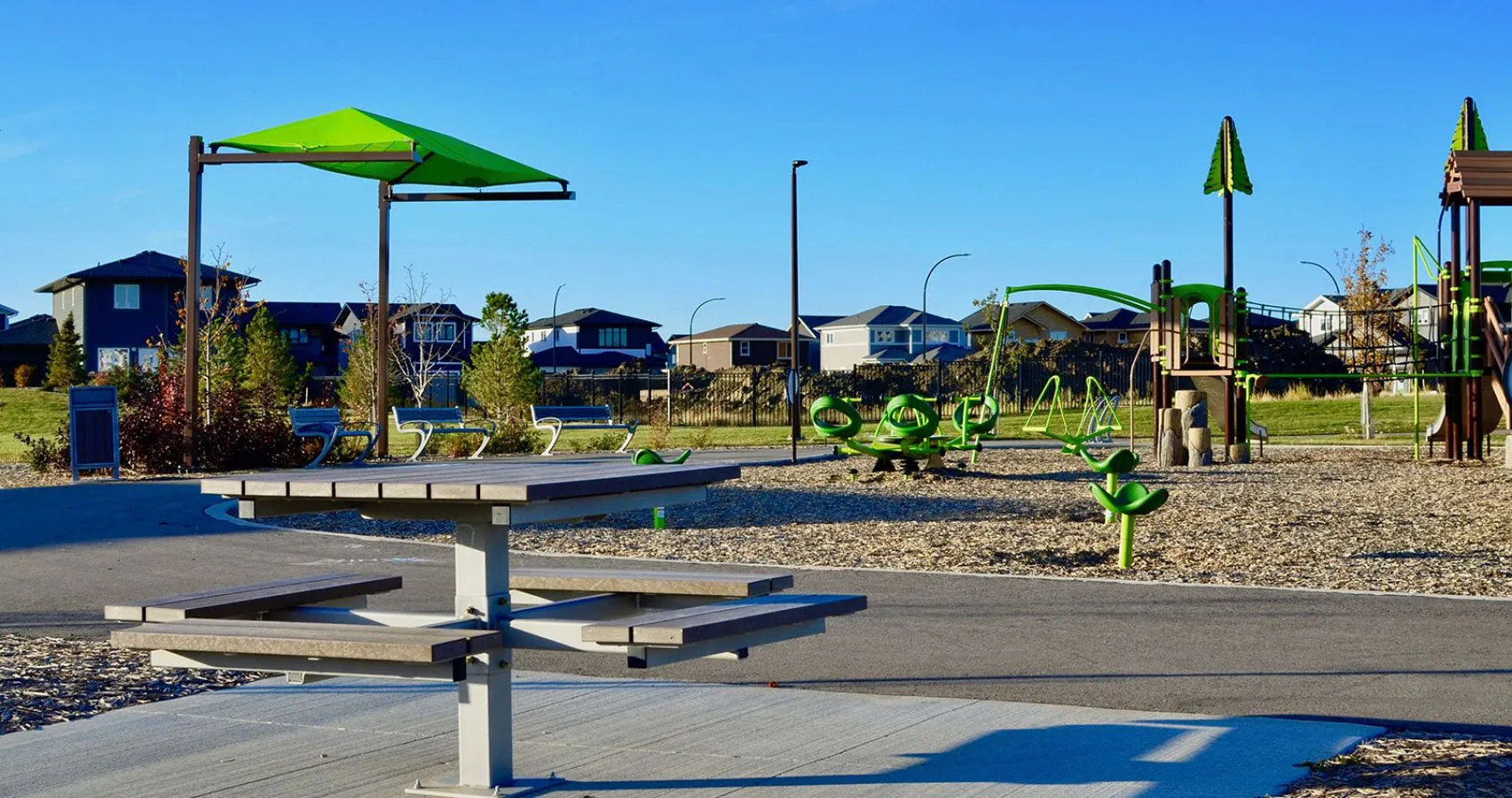 This single pedestal picnic table offers a comfortable and accessible space for outdoor dining and relaxation. The table is located in a park setting with playground equipment and residential houses in the background.