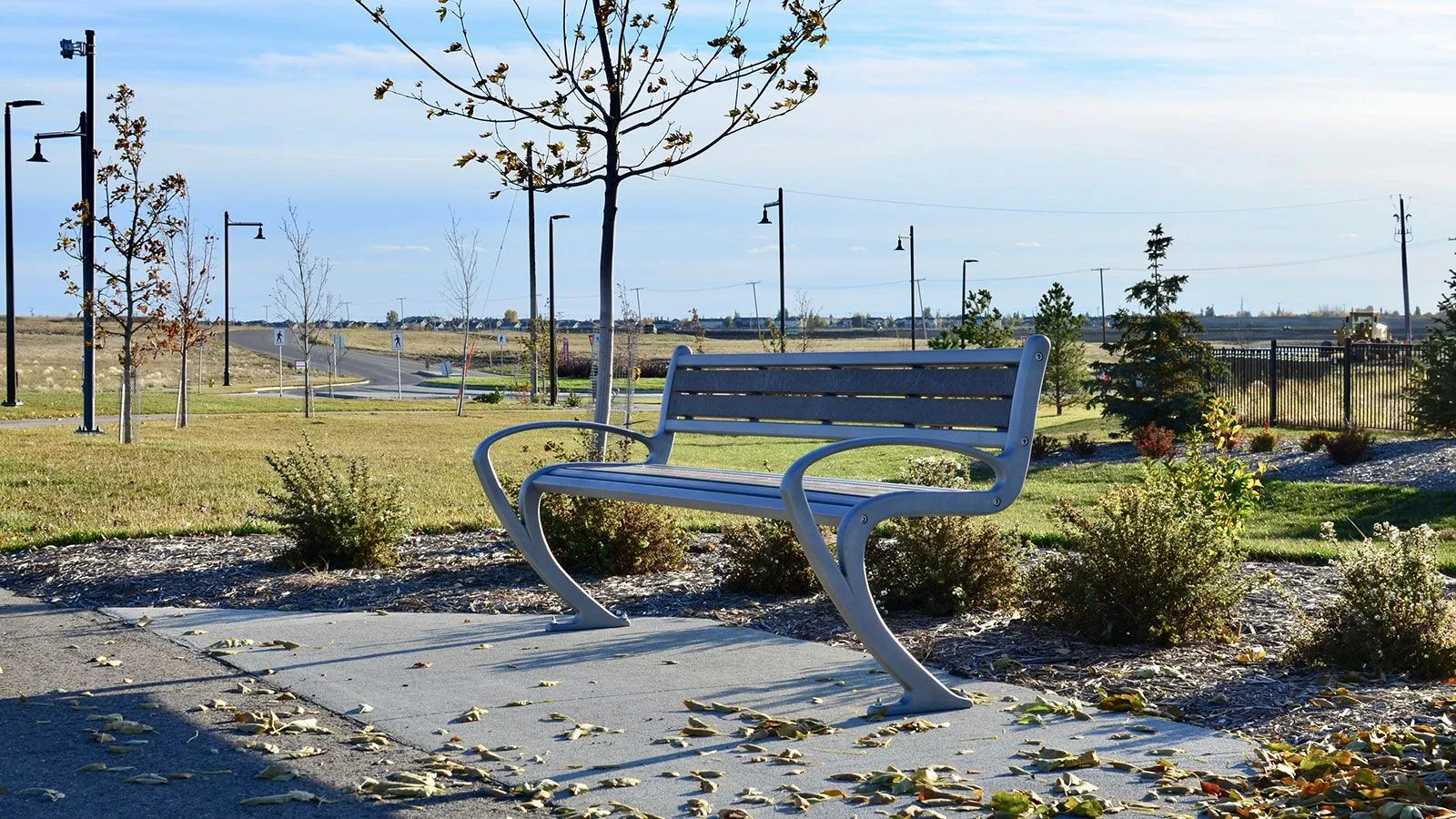 TENAJ bench with curved armrests in a park setting. The bench features recycled plastic lumber slats and a powder-coated metal frame.