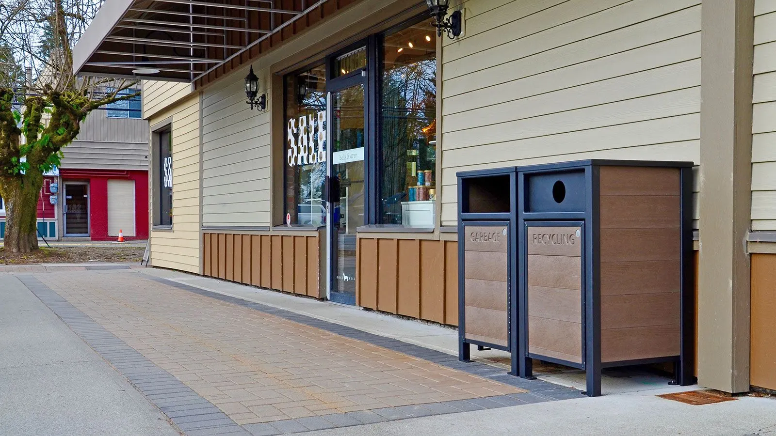 Dual garbage and recycling receptacles with recycled plastic lumber panels and a powder-coated metal frame provide a convenient waste management solution for commercial spaces. The receptacles are located outside a shop on a paved walkway.