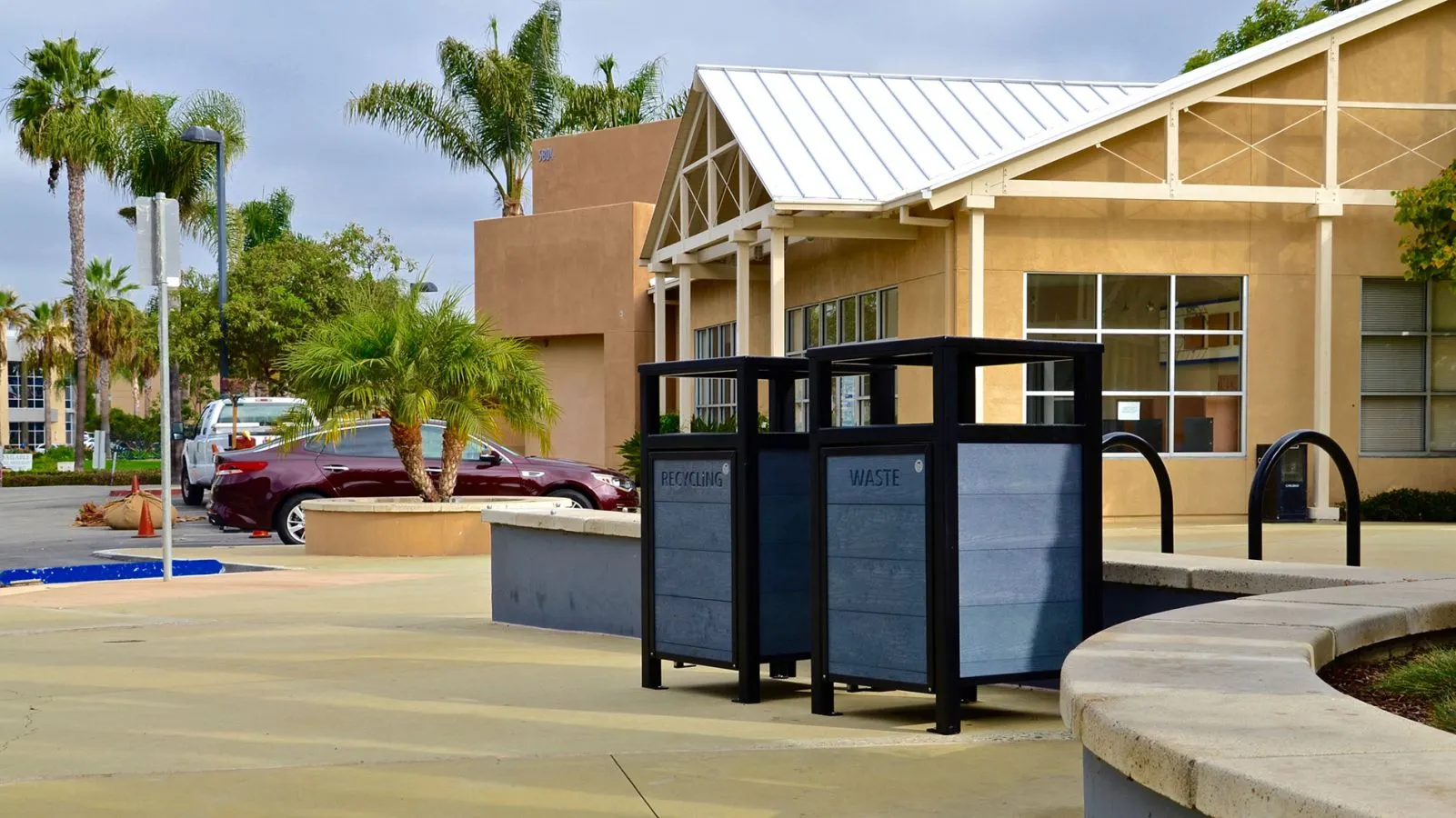 Dual waste and recycling receptacles with a modern design stand in an outdoor setting. The receptacles feature recycled plastic lumber panels and black powder-coated frames.