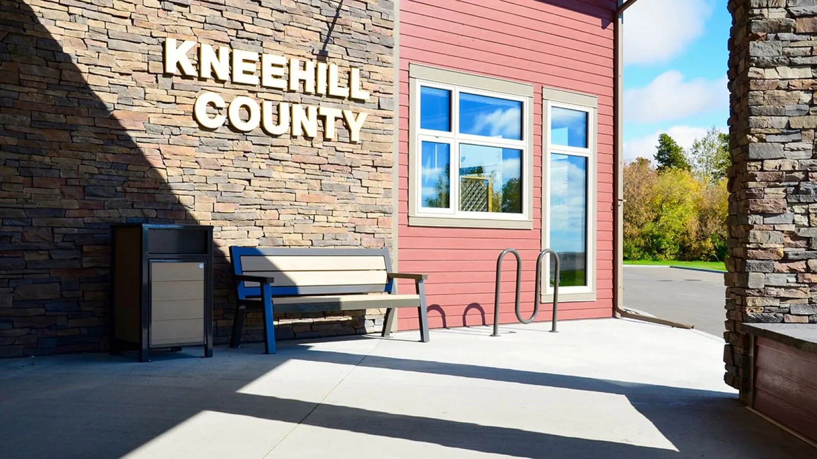Outdoor setting featuring a bench, bike rack, and waste receptacle. The site furniture complements the building's stone and siding exterior.
