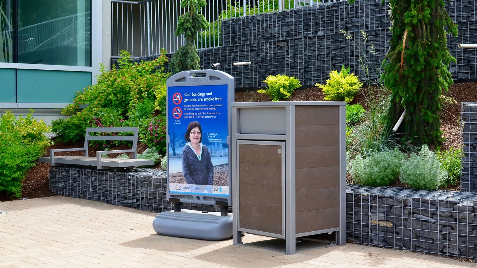 Urban Form waste receptacle with a brown recycled plastic lumber body and silver powder-coated frame. The bin is located outside a building with a smoke-free grounds sign.
