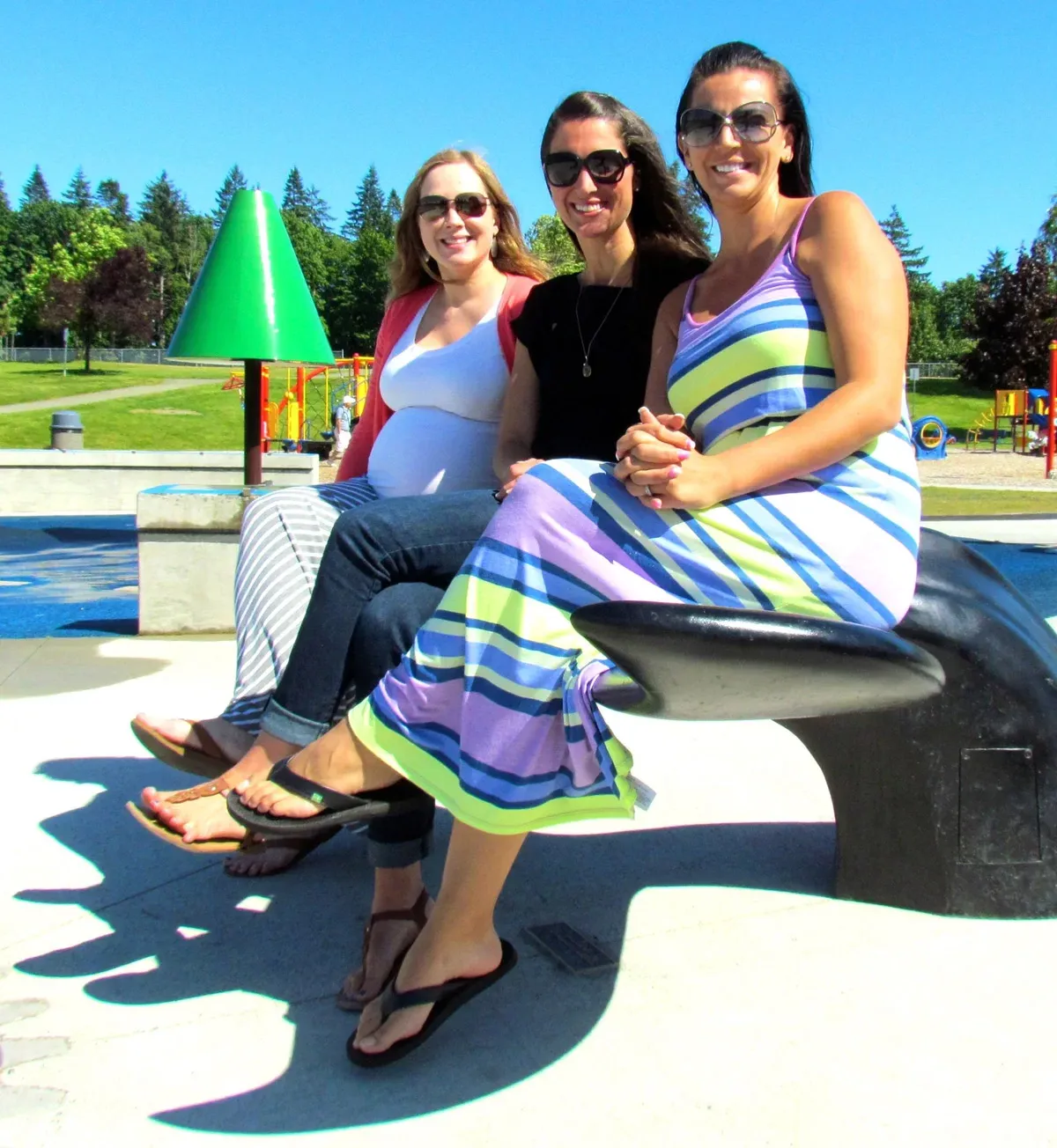 Three women are sitting on a playful whale tail bench in a park setting. The bench adds a whimsical touch to the outdoor space.