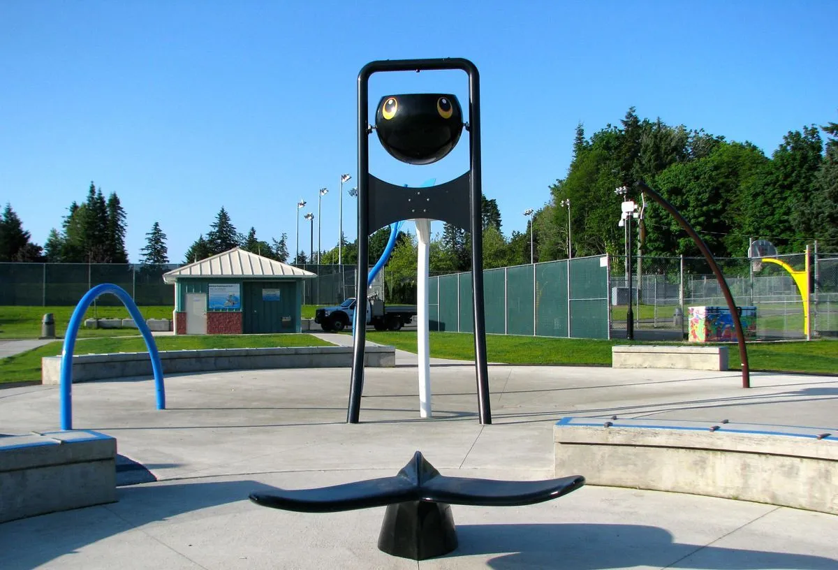 A whimsical whale tail bench and other play structures create a fun splash park environment. The black whale tail provides a unique seating option in this recreational space.