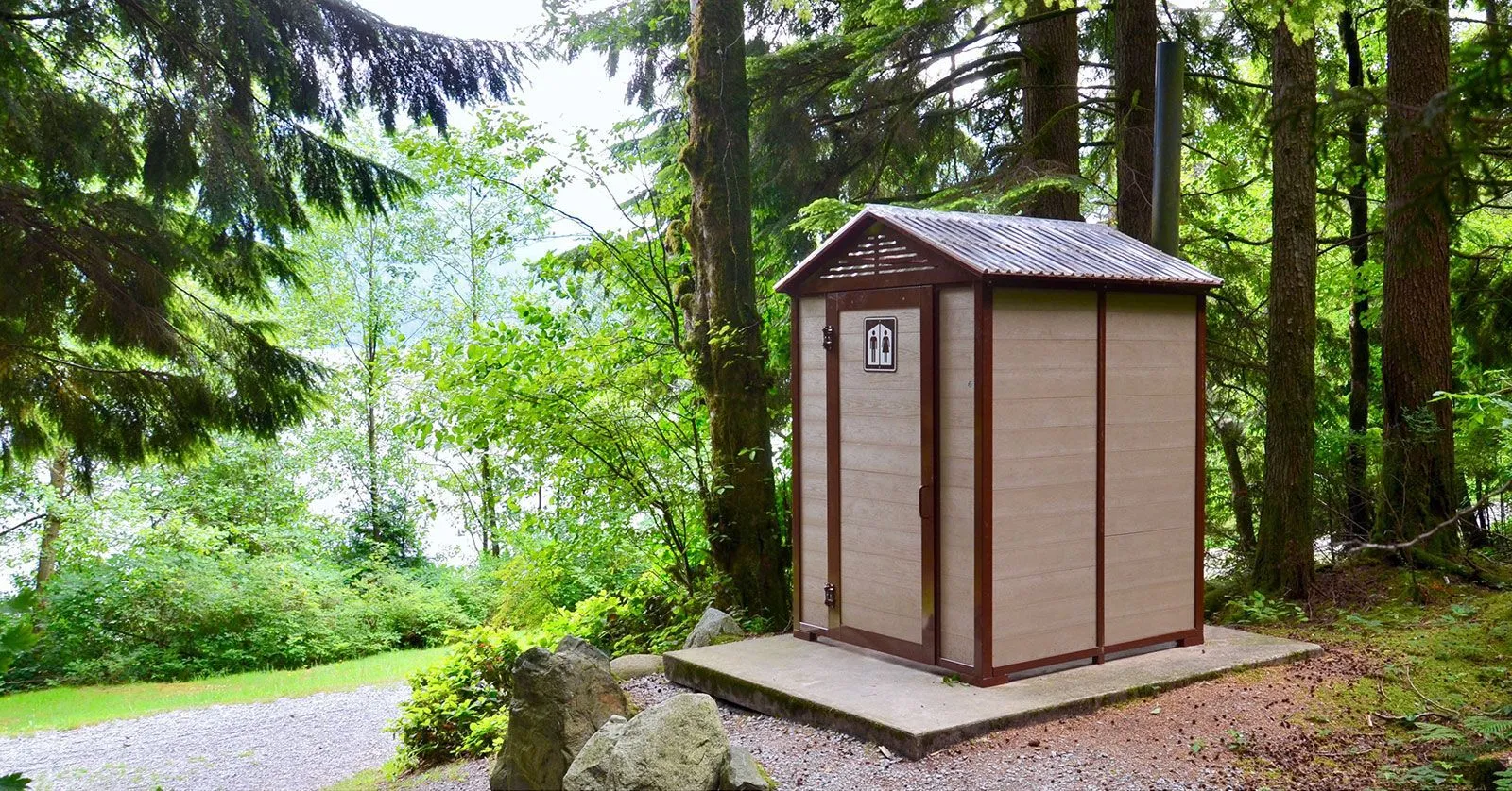 Wheelchair-accessible pit toilet building in a wooded park setting. The structure features recycled plastic lumber and dark trim.