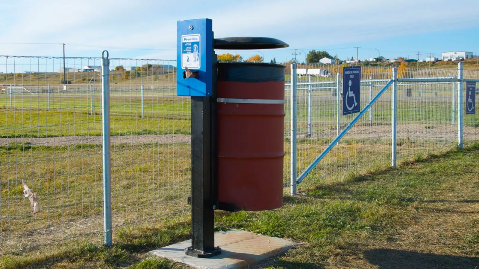 A Wrap 45 waste receptacle is installed at a dog park. The unit features a blue dispenser and a red barrel.