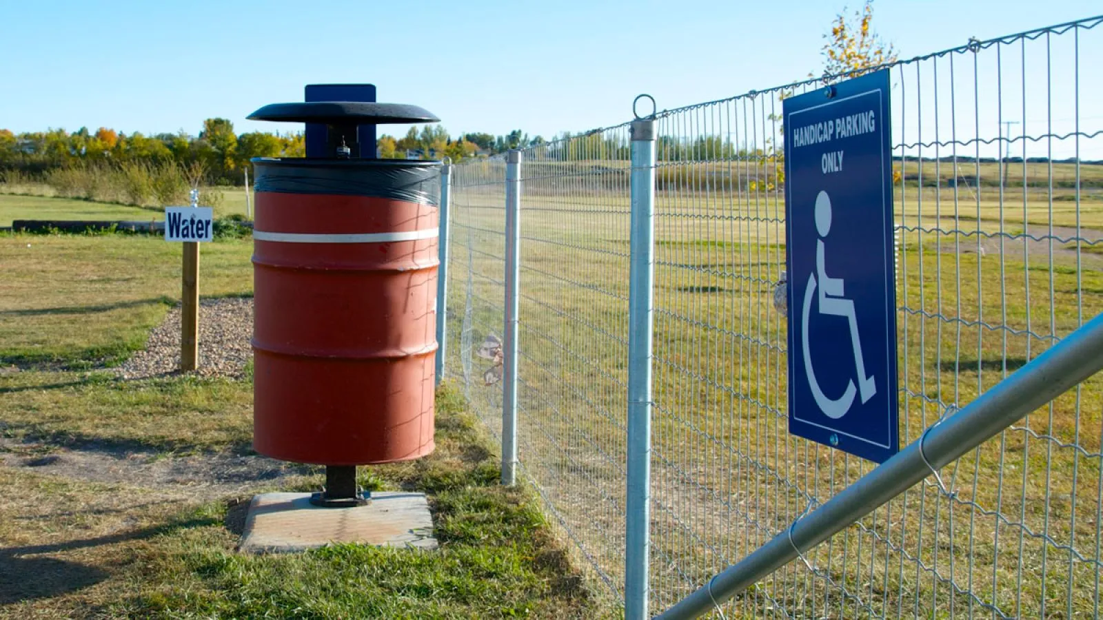 A red Wrap 45 waste receptacle stands near a fence in a park setting. The bin is made of a red barrel with a black lid.