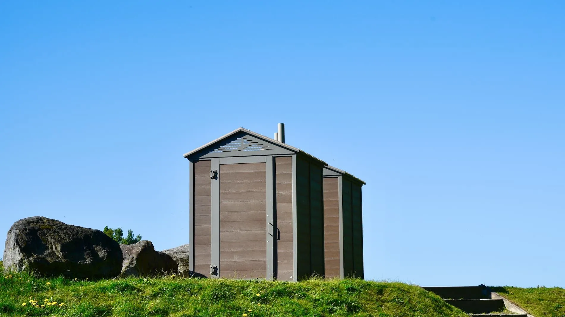 A pair of John Regular Outhouse Buildings stand on a grassy hill under a clear sky. These durable and functional structures provide essential facilities in outdoor settings.