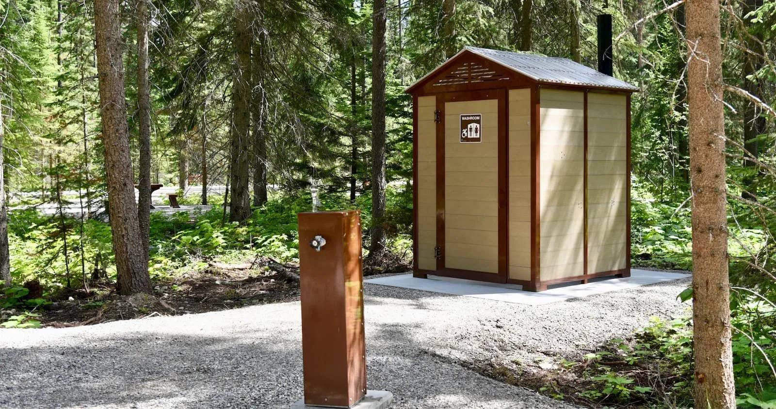 A Jumbo John outhouse building and a water fountain stand in a wooded area. The outhouse has a brown trim and beige panels, while the water fountain is a dark brown.
