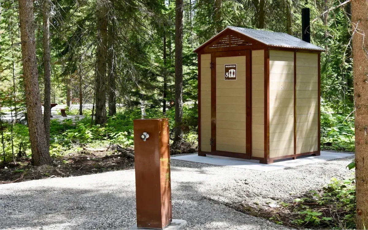 A brown water fountain stands near a washroom building in a wooded area. The washroom has light-colored siding with dark brown trim.