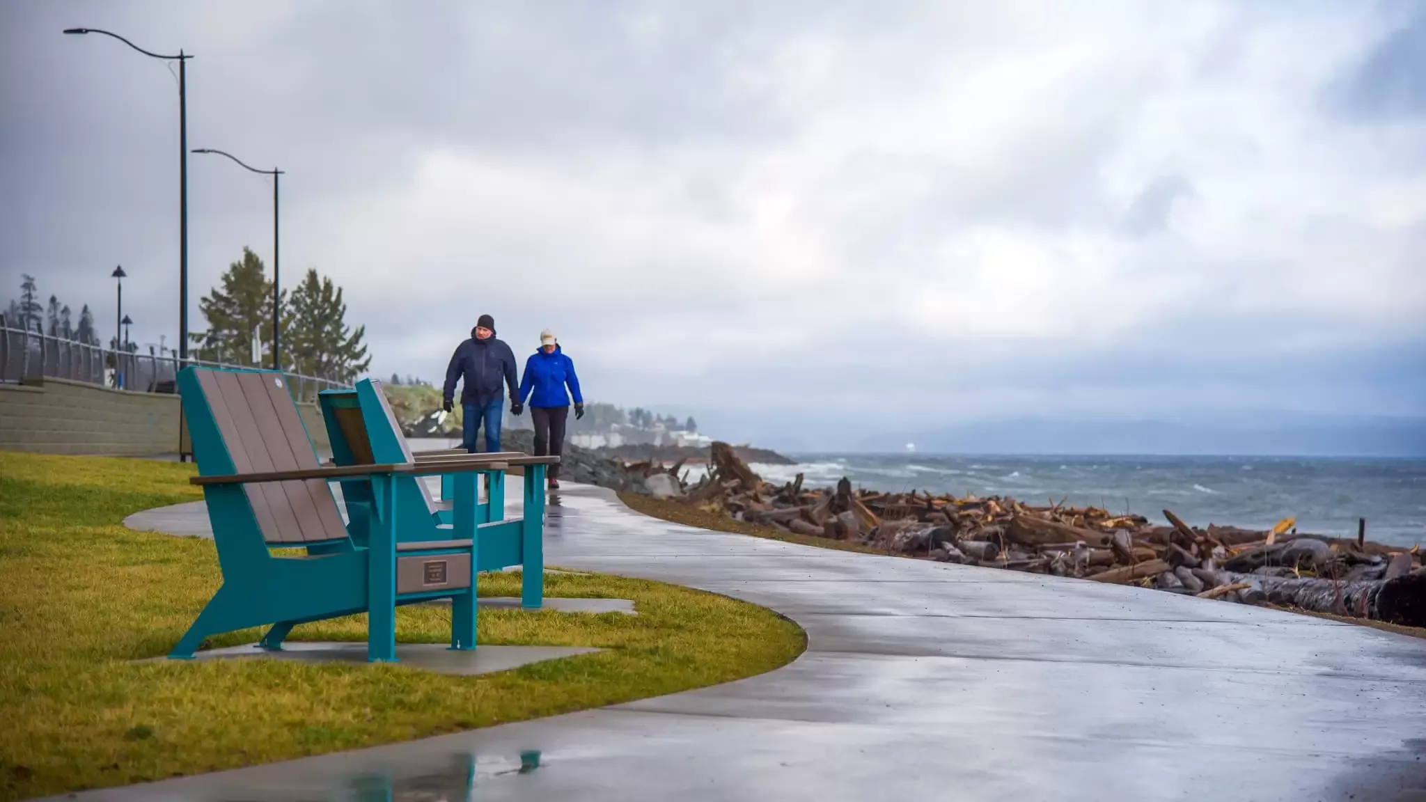 Two Adirondack-style lounge chairs face the ocean along a paved waterfront path. The chairs feature a teal frame and brown seat slats.