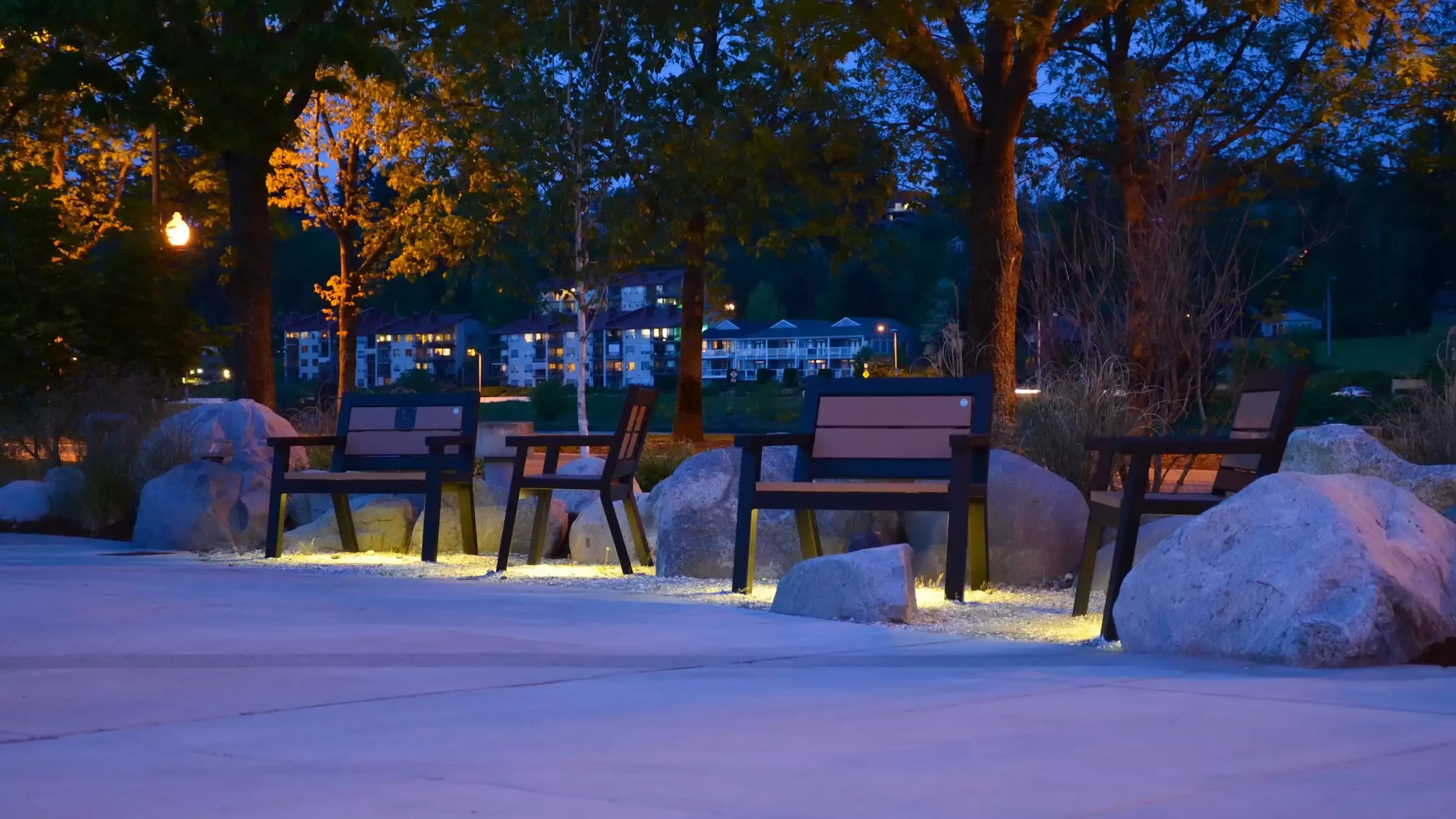 Rutherford benches line a walkway at night, illuminated by ground lighting. These benches feature recycled plastic lumber seats and backs with a dark powder-coated frame.