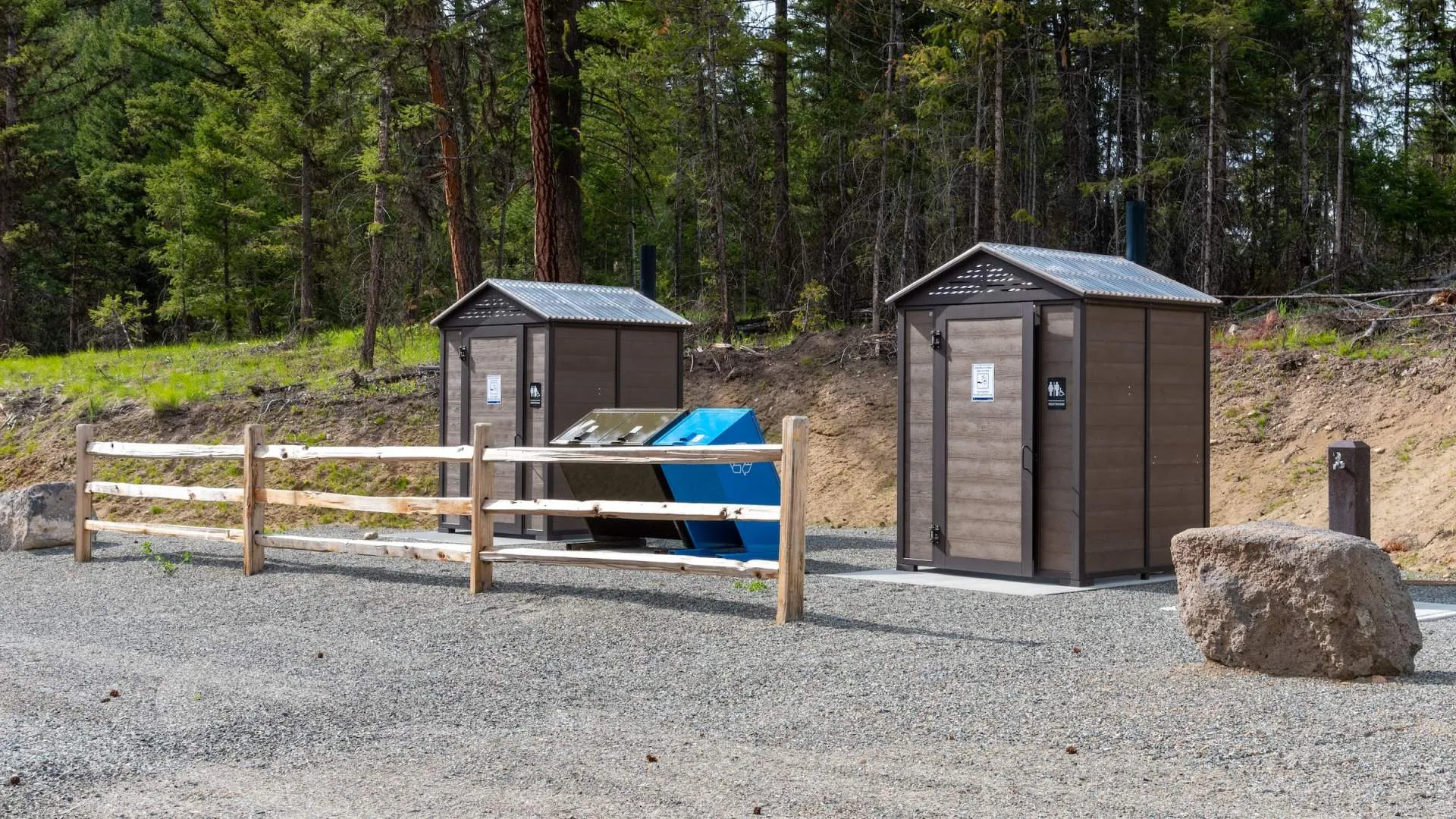 Two outhouses stand in a gravel area near a forest, with a split rail fence and recycling bins nearby. These buildings provide essential facilities in a natural park setting.