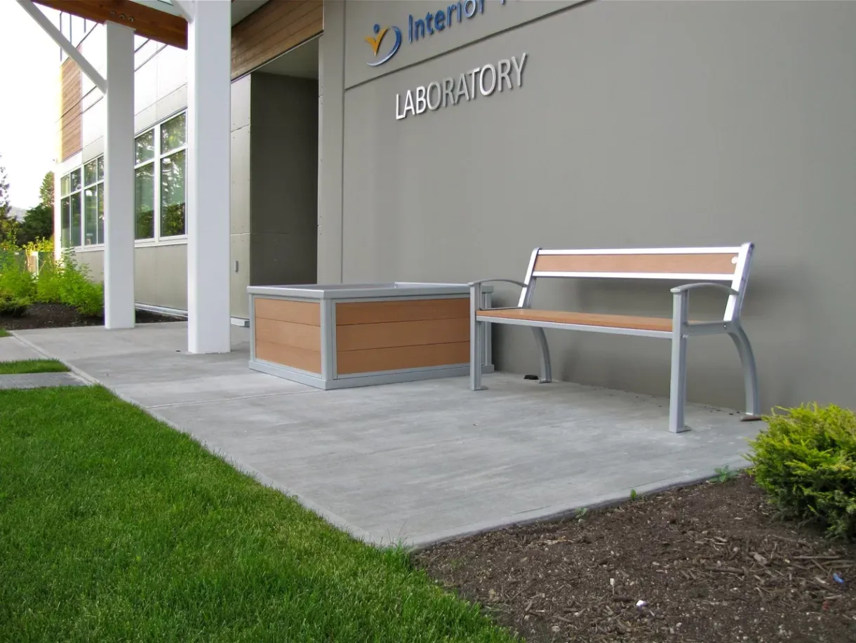 This outdoor setting features a Beselt bench paired with a square planter, creating a welcoming space outside a building. The bench and planter combine recycled plastic lumber with a powder-coated metal frame.