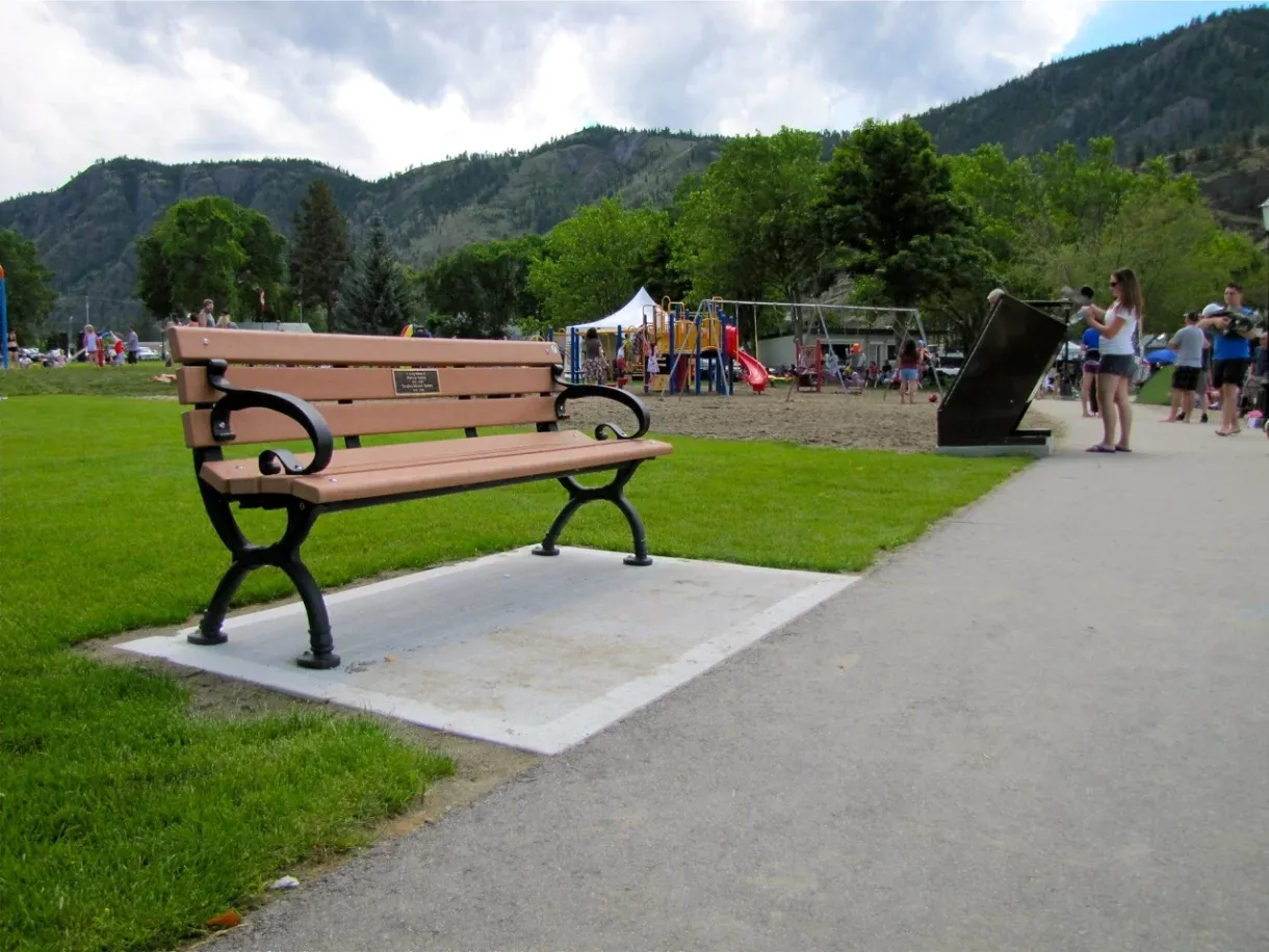 A Classic bench provides seating in a park setting, with a playground and mountains in the background. The bench features recycled plastic lumber and a black powder-coated frame.