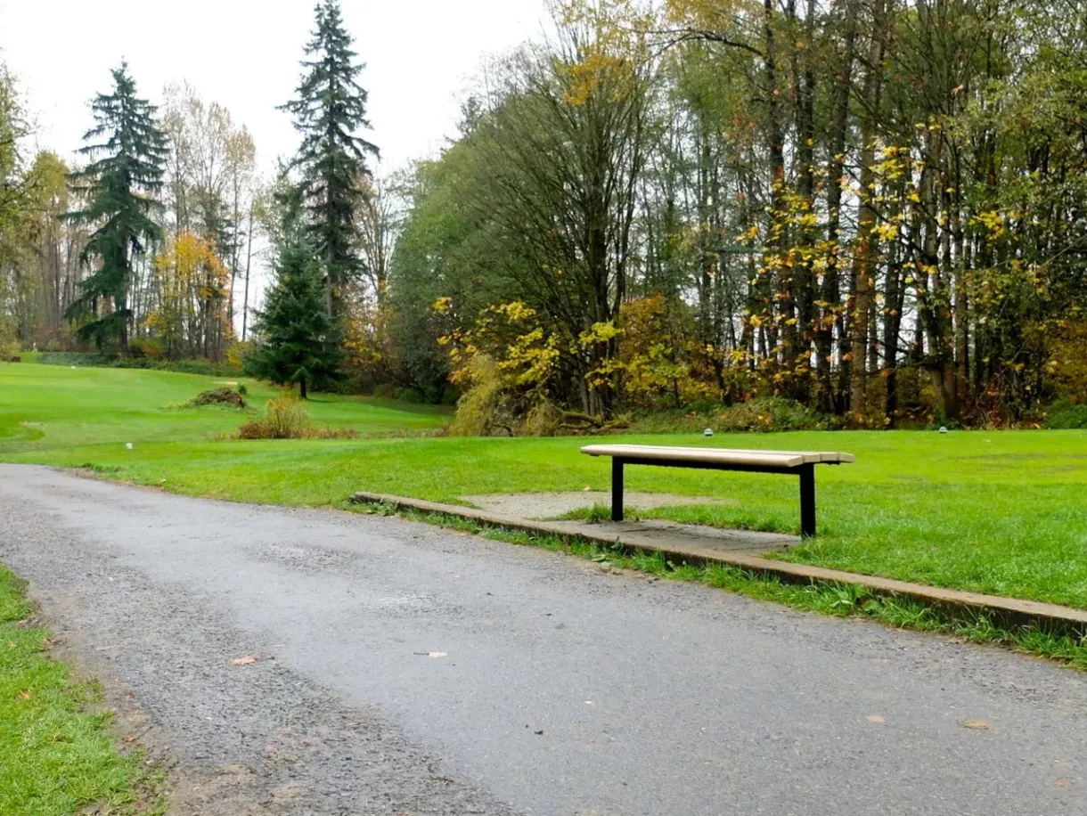 Contoured Backless Bench provides simple seating along a paved path at Riverway Golf Course in Burnaby, British Columbia. The bench features a light-colored lumber seat and black metal frame.