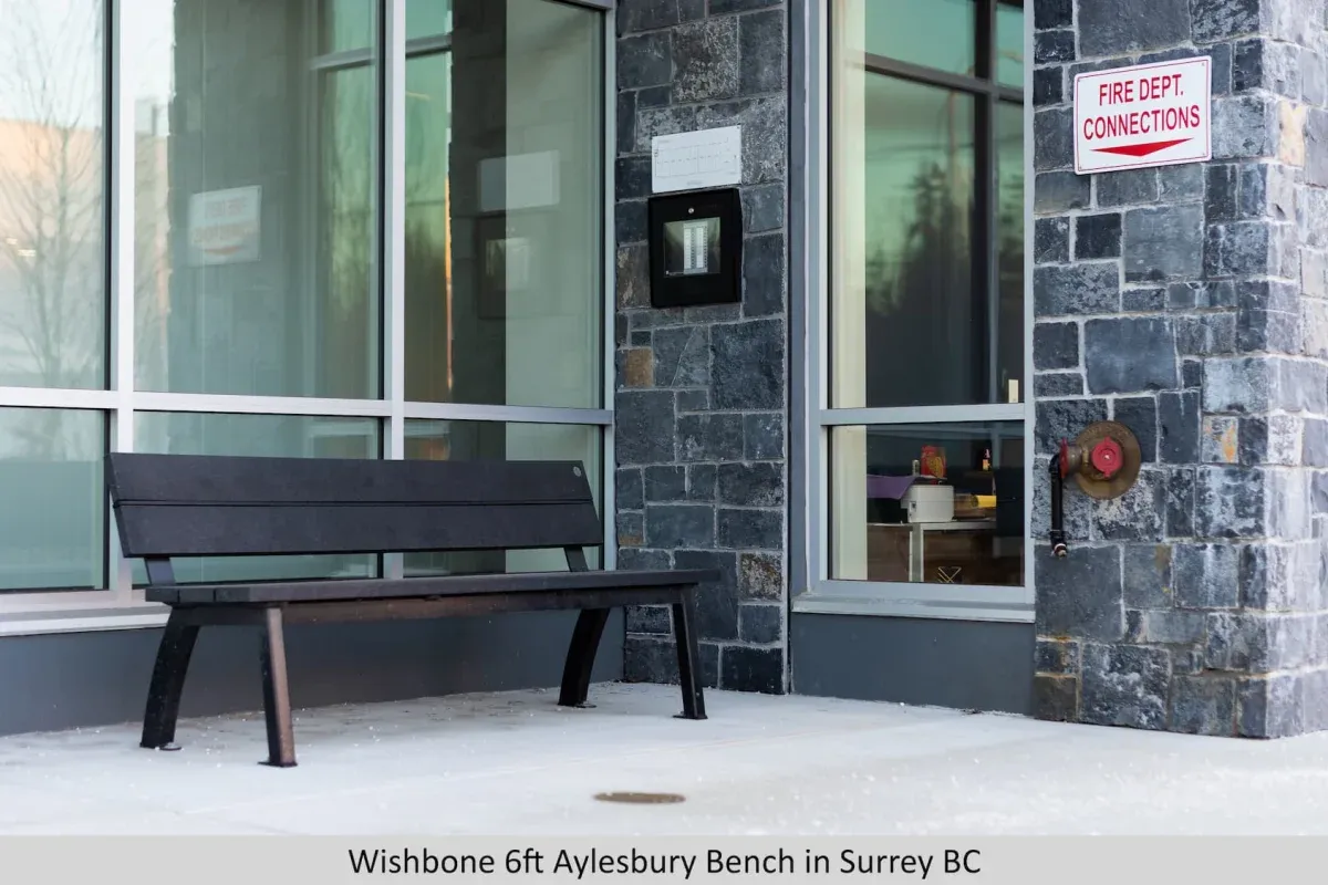 A black Aylesbury bench sits outside a building with large windows and a stone facade in Surrey, British Columbia. The bench provides seating near a fire department connection point.