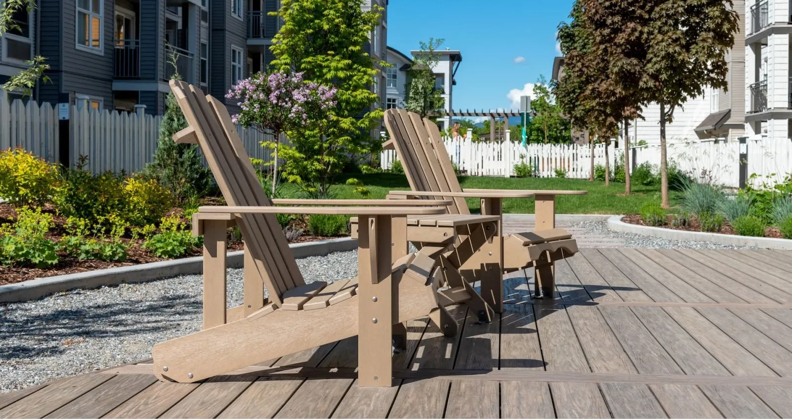Two Adirondack chairs and a small table made from recycled plastic lumber sit on a wooden deck in a landscaped outdoor area. The setting features modern residential buildings and manicured gardens.
