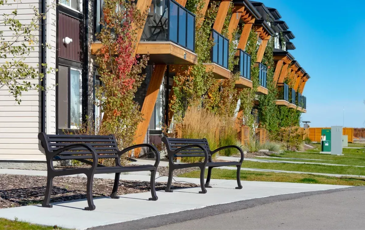 Two Mountain Classic benches provide seating along a paved walkway in front of a modern building with vine-covered balconies. The benches offer a place to rest and enjoy the surrounding landscape.