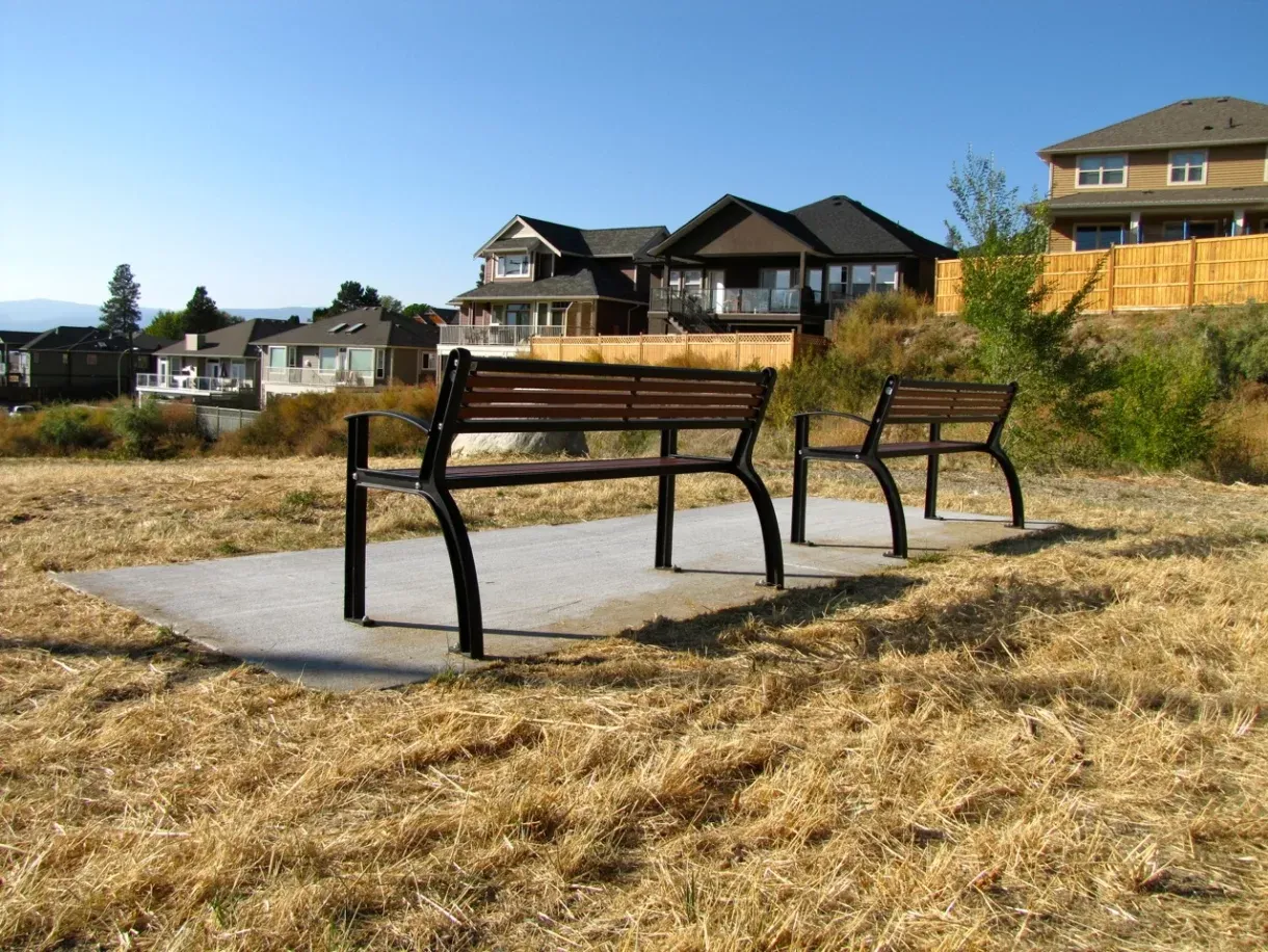 Two metal benches with recycled plastic lumber slats sit on a concrete pad in a grassy area. Residential houses are visible in the background.
