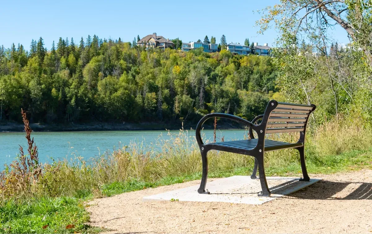 A Mountain Classic bench sits along a riverside path, offering a place to rest and enjoy the view. The bench features recycled plastic lumber and a durable powder-coated metal frame.