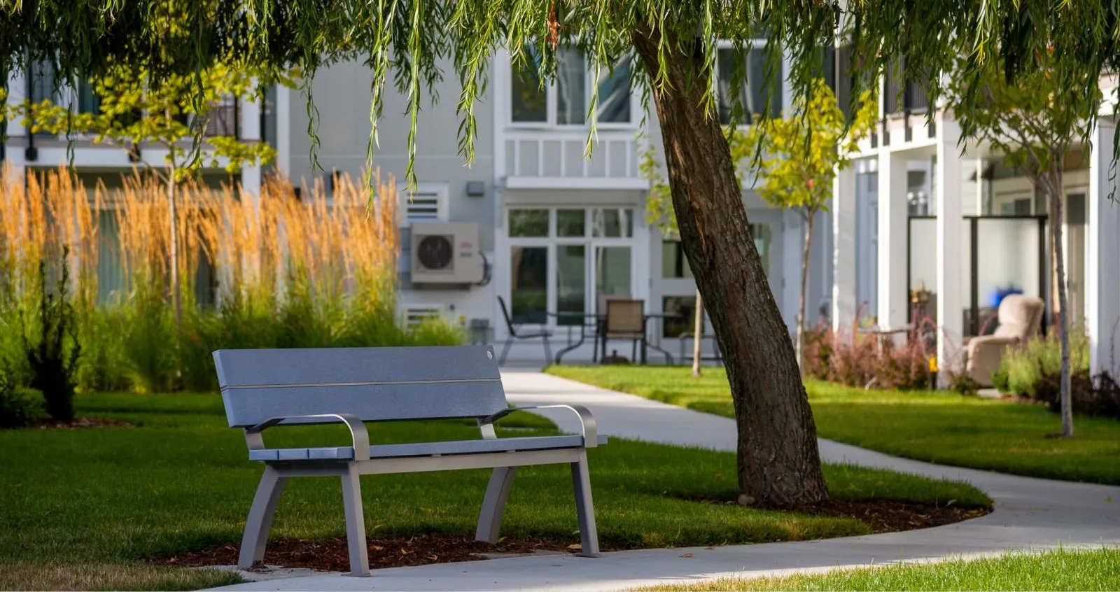 An Aylesbury bench with armrests sits on a pathway surrounded by green grass and landscaping. The bench provides a place to rest and enjoy the outdoor environment.