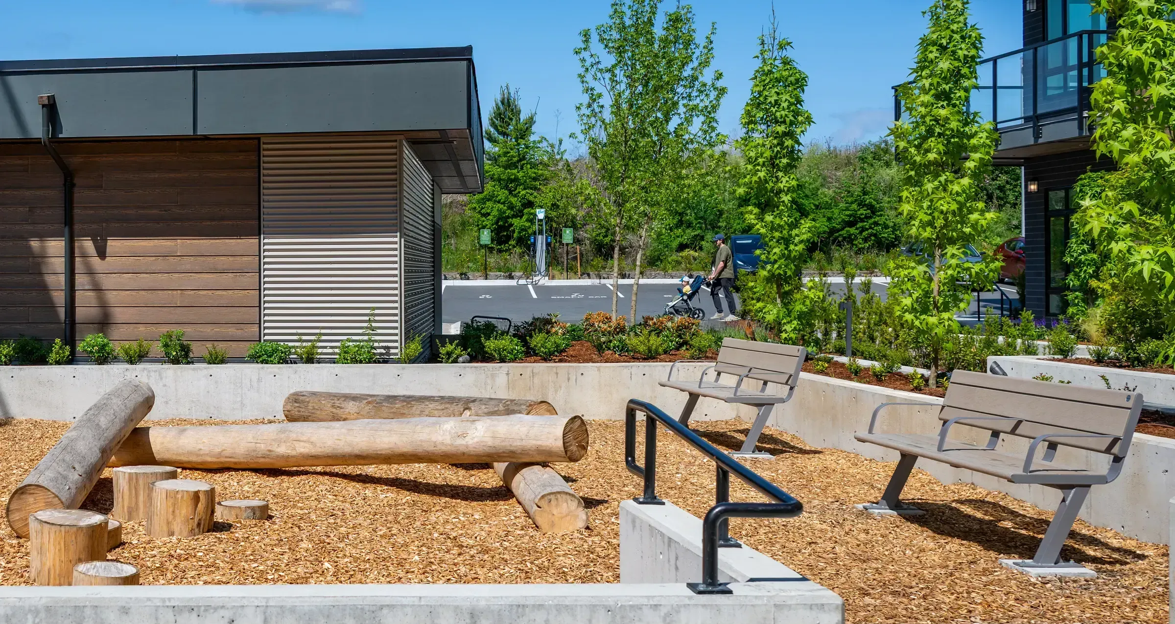 Bayside benches provide seating in a landscaped outdoor area at Bella Vista Apartments. The setting features a natural playground with logs and wood chips.