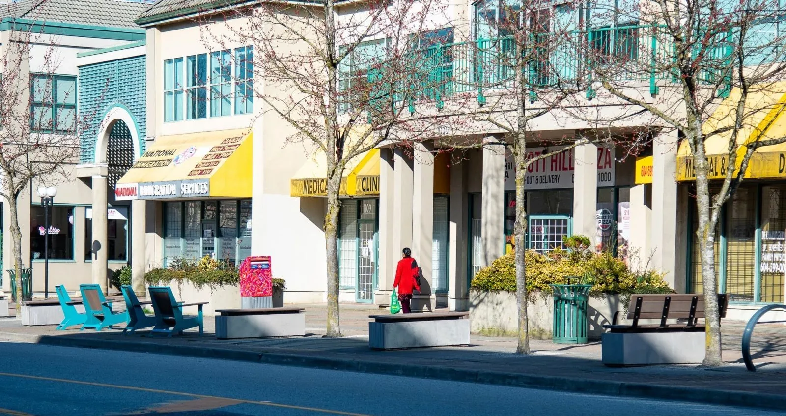Streetscape featuring backless benches with concrete bases and timber tops, and a green waste receptacle. The benches provide seating along a commercial street with shops and businesses.