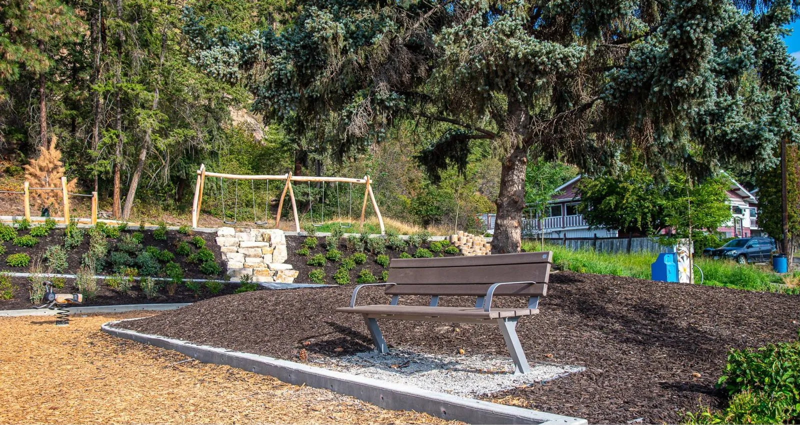 A Bayview bench sits in Ballou Park, Kelowna, BC. The bench features recycled plastic lumber seat slats and a metal frame, providing a comfortable place to rest near the playground.