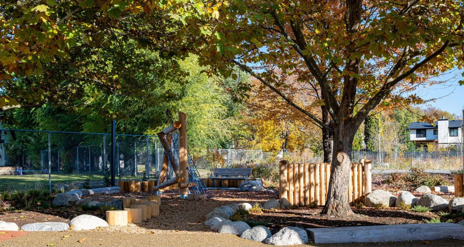 A natural playground features a Bayview Bench nestled among trees and play structures. The setting includes wood chip ground cover, rocks, and wooden elements.