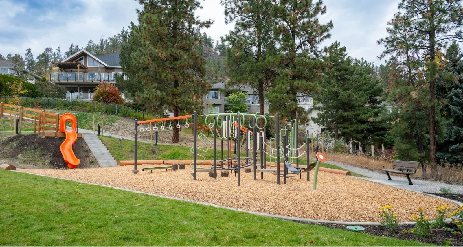 A park playground features a Bayview bench near a play structure. The setting includes trees, grass, and residential buildings in the background.