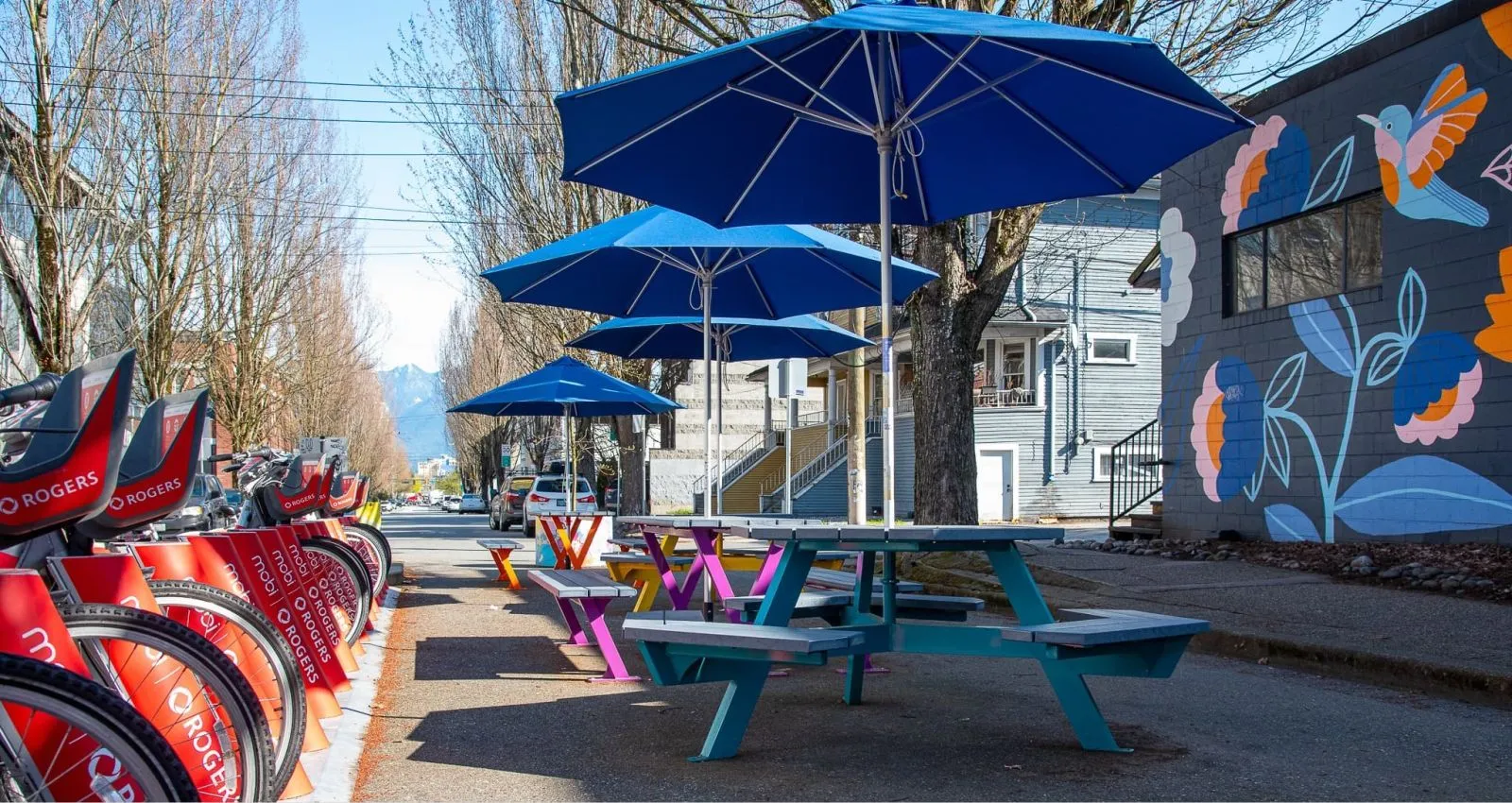 Colorful hexagonal picnic tables create a vibrant outdoor dining space. The tables feature a mix of powder-coated metal frames and recycled plastic lumber tops, complemented by blue umbrellas and a mural in the background.