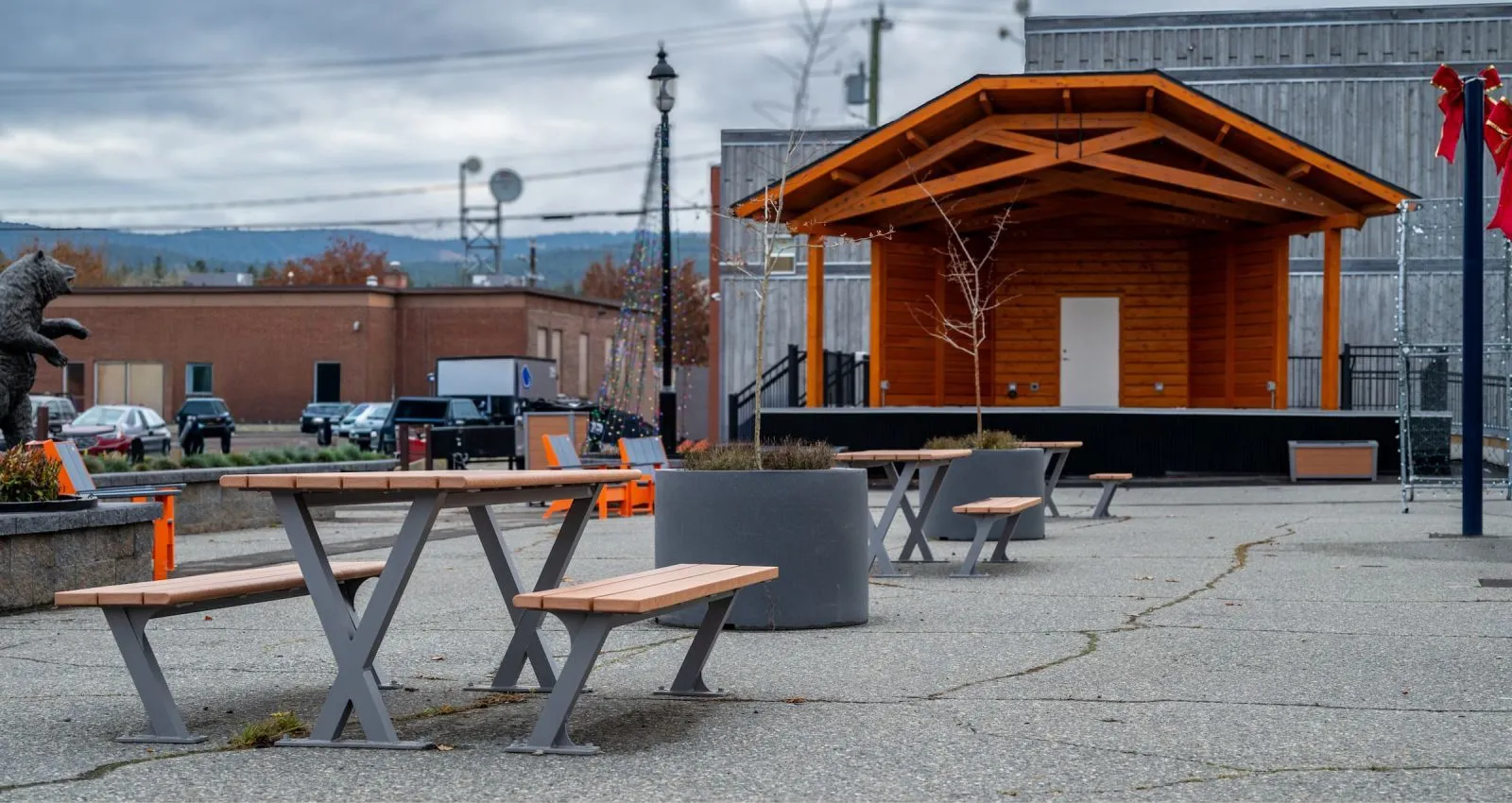 Bayview picnic tables provide outdoor seating in a public square. The tables feature recycled plastic lumber and powder-coated metal frames, complemented by concrete planters and a wooden stage in the background.