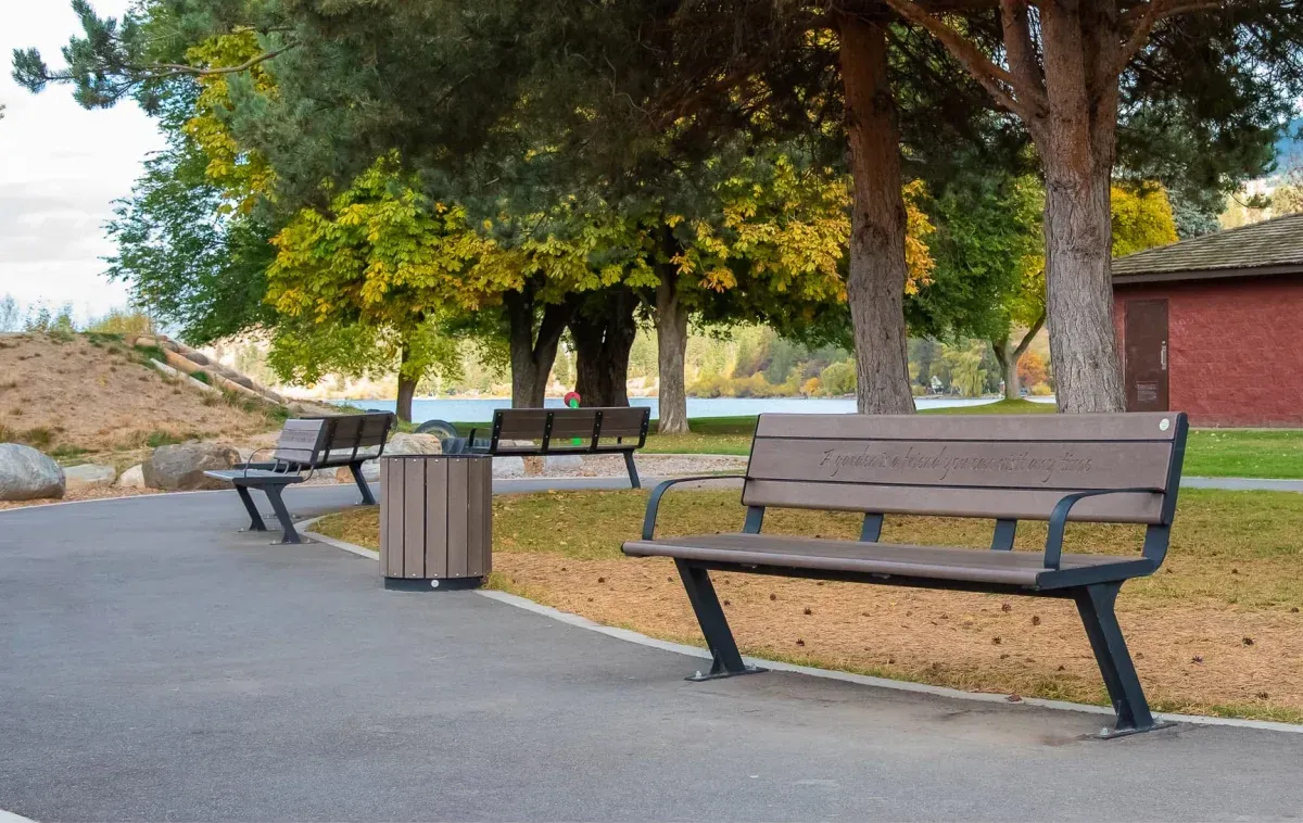 Bayview Plus benches and a waste receptacle line a paved path in Beasley Park. The park features mature trees and a lake in the background, creating a serene outdoor space.