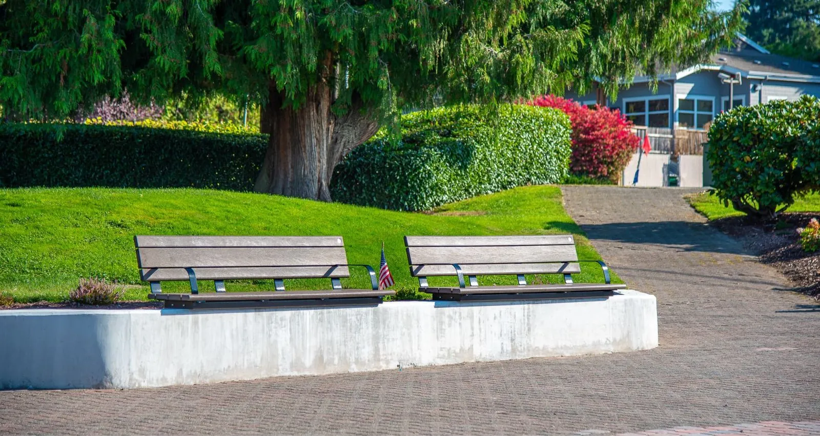 Two wall-mounted benches with recycled plastic lumber seating are installed along a concrete wall in a park-like setting. The benches provide a place to rest and enjoy the surrounding greenery.