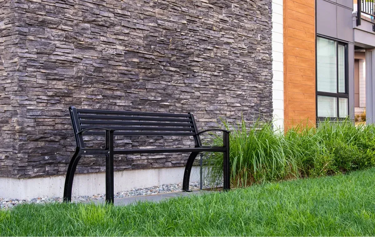 A black Beselt bench sits on a grassy area next to a building with a stone facade. The bench offers seating in an outdoor space.