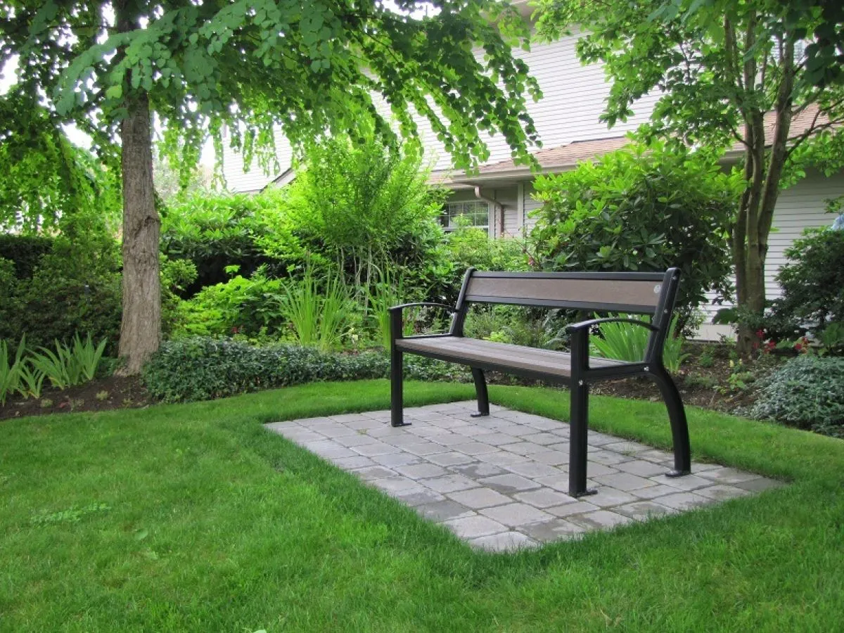 A Beselt bench sits on a paved area surrounded by lush greenery. This durable bench provides comfortable seating in outdoor spaces.