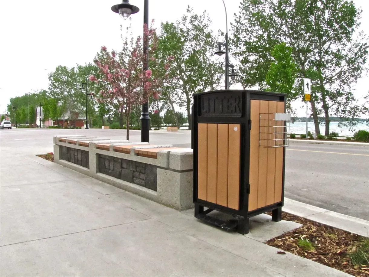 Beselt square top waste receptacle with a black frame and sand-colored recycled plastic lumber stands on a paved area next to a concrete bench in Sylvan Lake, Alberta. The litter bin features a metal cage for holding pamphlets or bags.